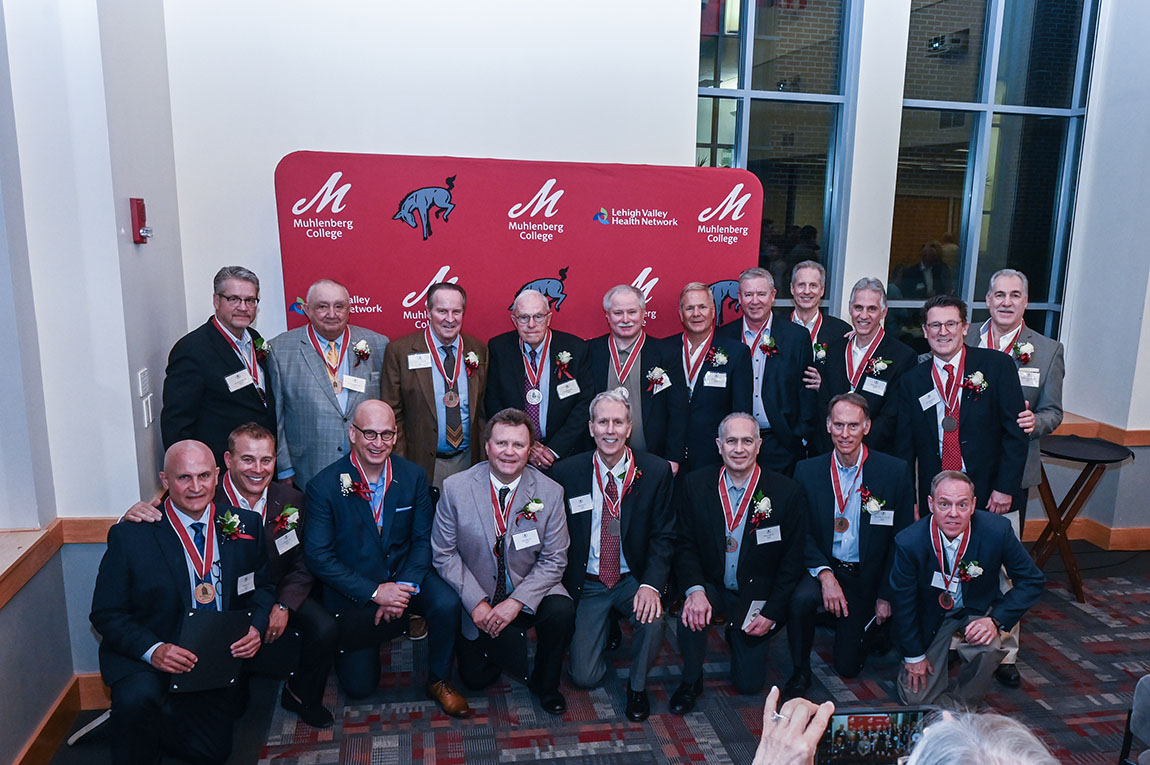 A large group of older men with medals on smile in front of a red backdrop